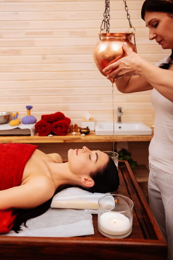 Therapist preparing Shirodhara — continuous medicated oil stream on forehead — for a resting patient.