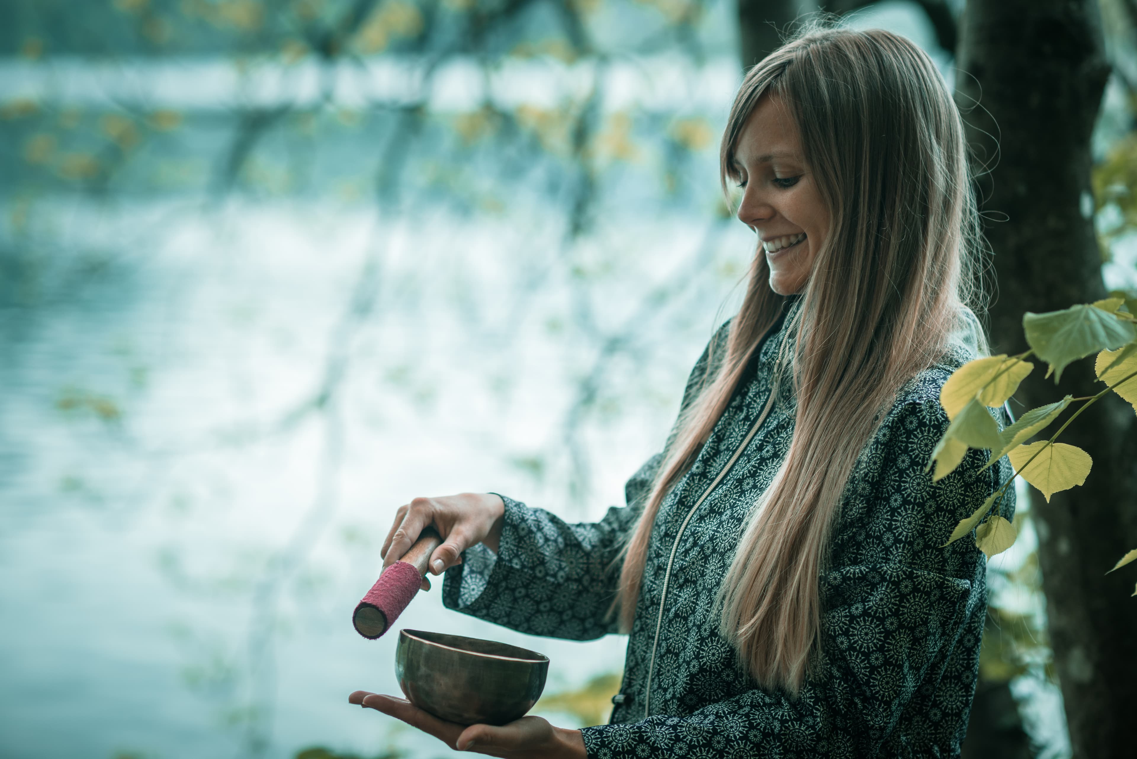 A practitioner gently using a singing bowl during a sound-healing session by a lake.