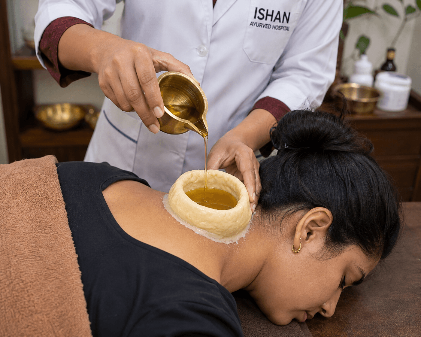 Therapist at Ishan Ayurvedic Hospital pouring warm oil into a dough ring sealed over a patient's neck.