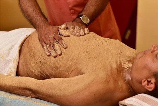 Therapist working coarse herbal powder across a patient's torso with firm upward strokes.